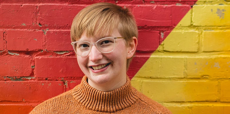 Person with short ginger hair standing against a red and yellow wall, smiling