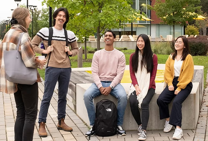 Five students chatting and sitting together outdoors on campus.