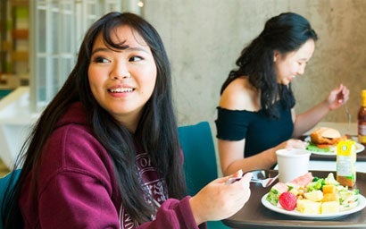 Two students eating in residence. One has a fruit salad and the other has a friend chicken sandwich.