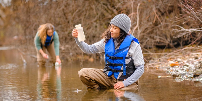 Two students in a creek, wearing hip waders and lifejackets, collecting water samples