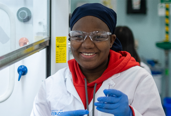 Person in a lab coat and safety goggles holding a small test tube inside a laboratory.