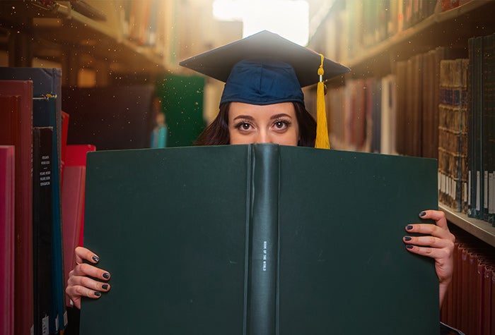 Student wearing a graduation cap, holding a book in front of them. Tall shelves lined with books are on either side of them.