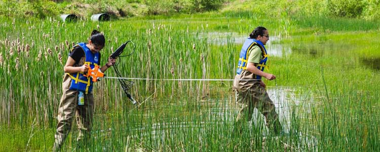Two students wearing life jackets and chest waders walking through water with a measuring device in hand