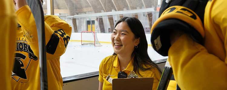 Studnent holding clip board. Hockey players on either side. Hockey rink in the background.