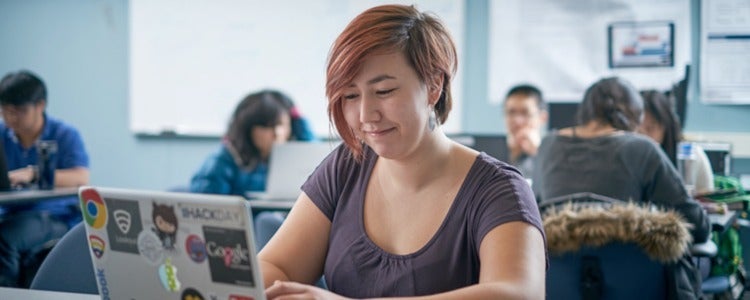 Student using laptop on desk with other students in background