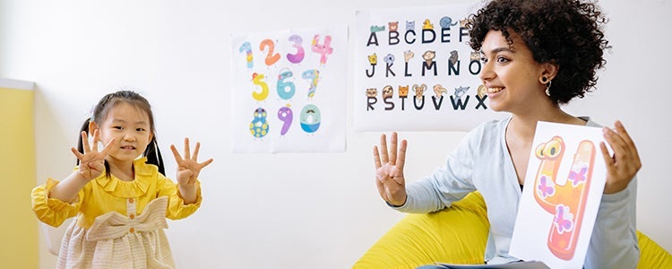  A teacher holding up a book with numbers on it. A young student to the left of her counting with her fingers.