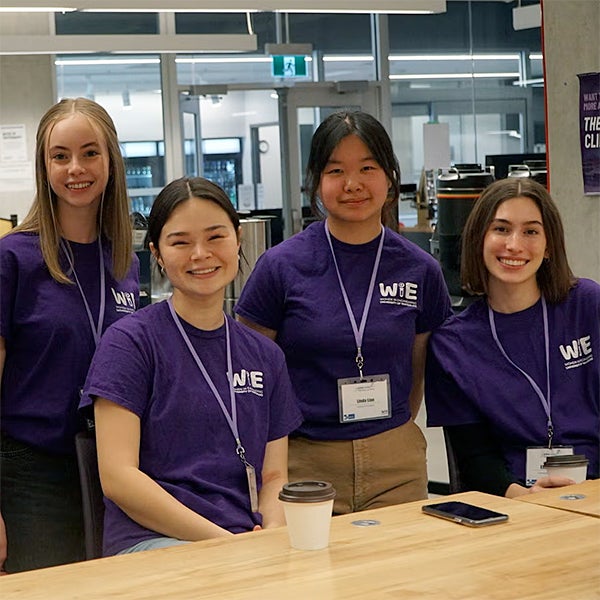 Four students in purple Women in Engineering shirts and name tags smile