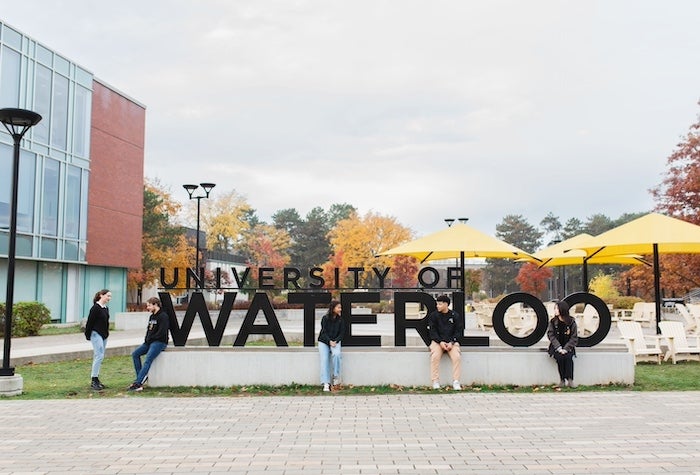 Students sitting in front of the University of Waterloo sign on campus.