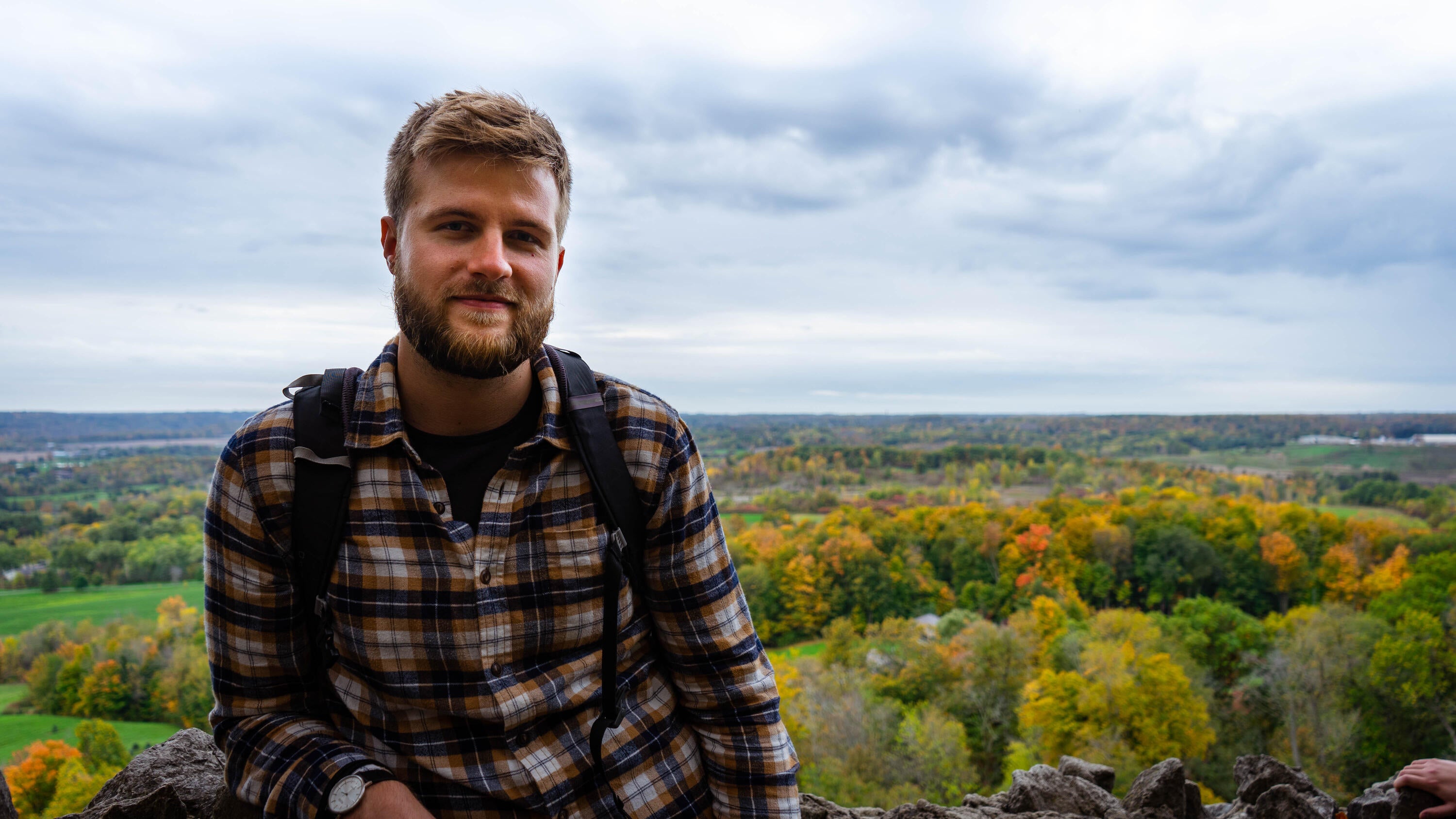 Upper body of Marvin Pafla in a checkered shirt with a backpack on standing in front of a forest 