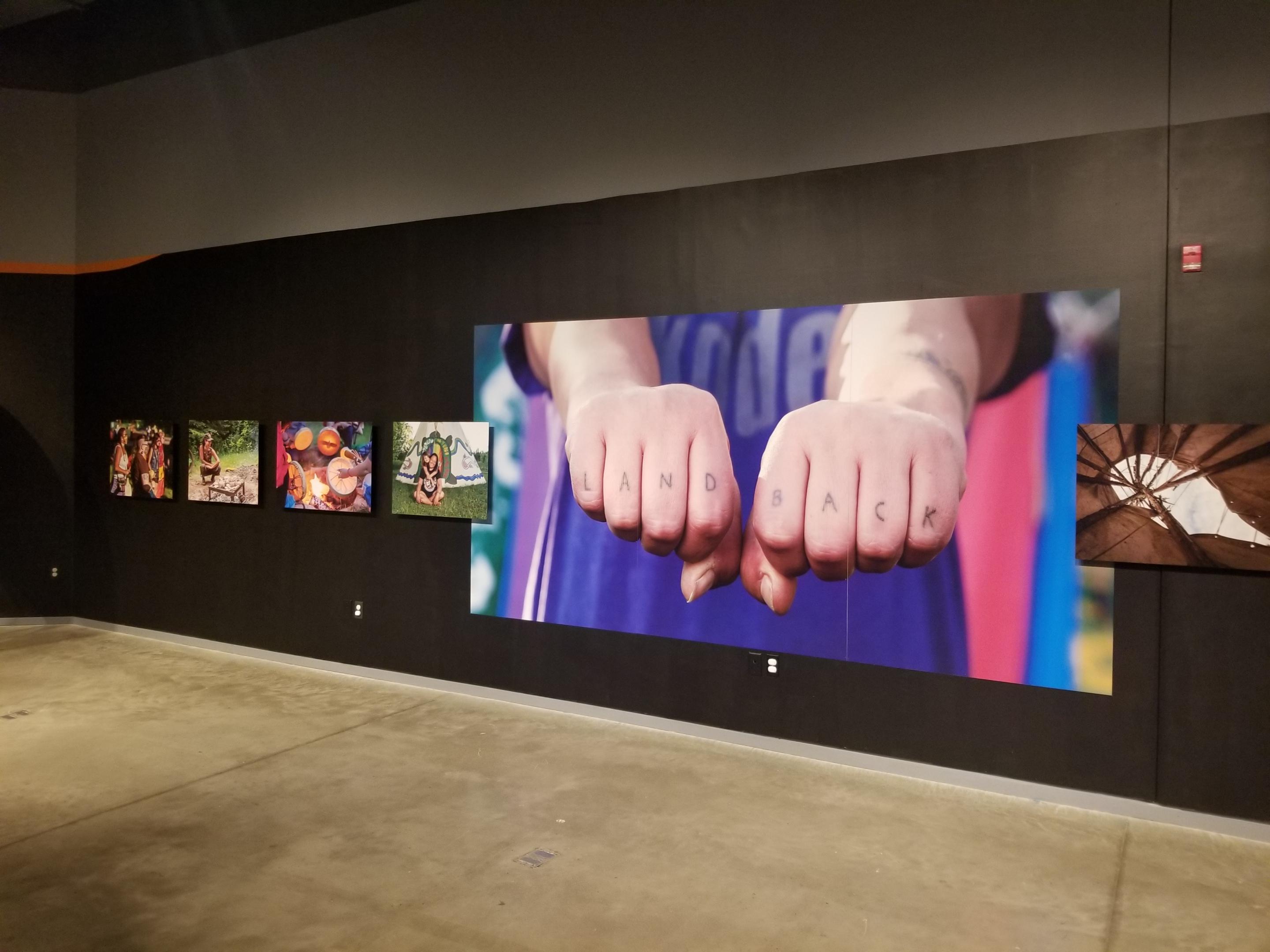 A series of photographs on a wall as part of the exhibition. The largest photo features two fists with LAND BACK written across the knuckles