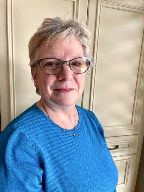 Anne is a middle-aged white woman with short white-blonde hair. She is wearing glasses and a blue shirt.