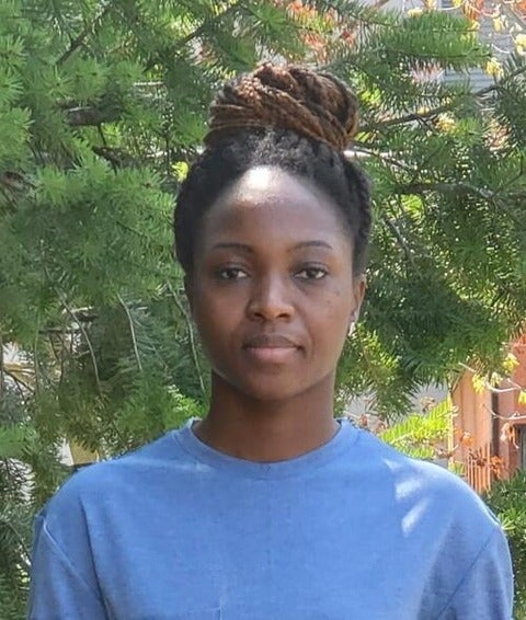 Headshot of Thelma Abu. Her hair is in a braided bun, she is wearing a blue T-shirt. Behind her is an evergreen tree.