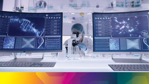 A person in a labcoat and protective gear looking through a microscope flanked by two computers showing data