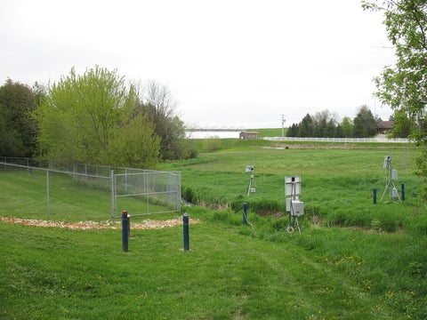 A grassy landscape within the Alder Creek watershed with groundwater monitoring instrumentation in the foreground, a chain-link fence, and a lake visible in the distance under a cloudy sky.