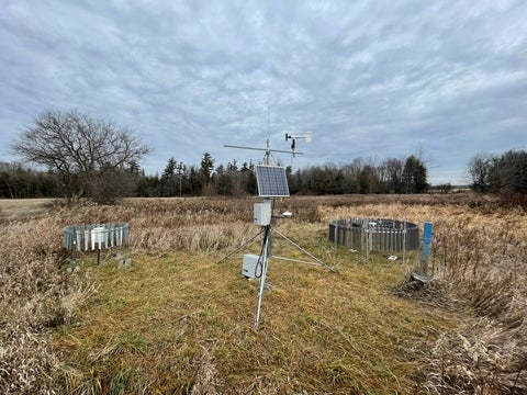 A monitoring station with various instruments and a solar panel stands in a grassy field in the Alder Creek watershed under a cloudy sky.