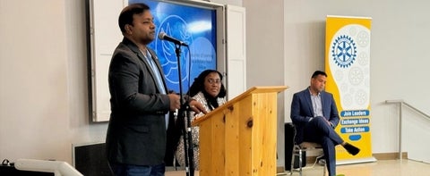 a man speaking at a podium, with two people sat on stage behind him listening