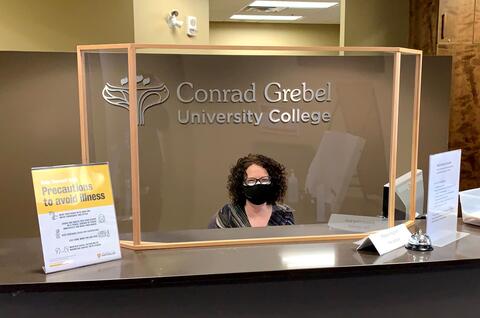Leanne sits at the Grebel front desk behind a plexiglass barrier