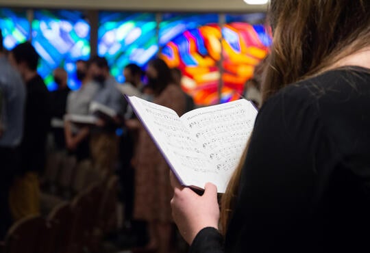 Person holding a choir book in a chapel with stained glass windows
