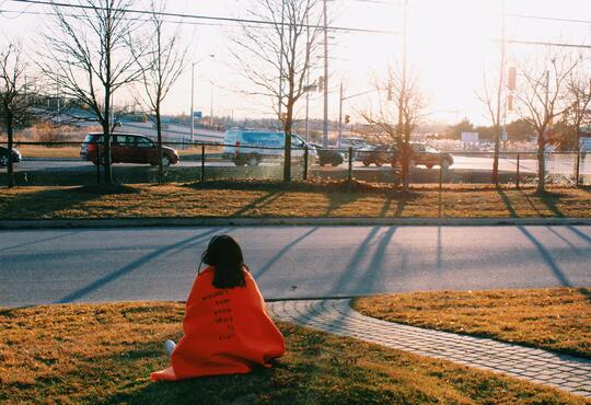 The artist sits outside on a front lawn by a road, wrapped in an orange blanket that reads 