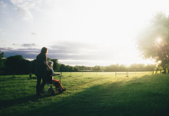 An elderly woman and caregiver enjoy a sunset in a green field