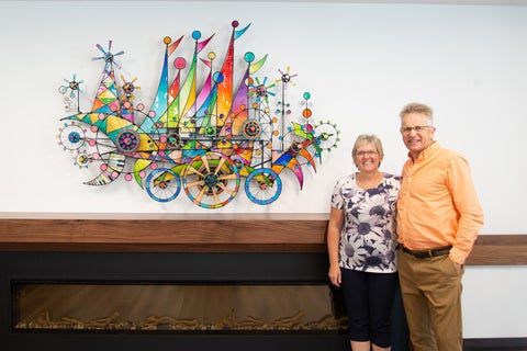Jim stands with his wife beside the colourful metal and resin scuplture, now hanging above the fireplace in the grebel dining room.