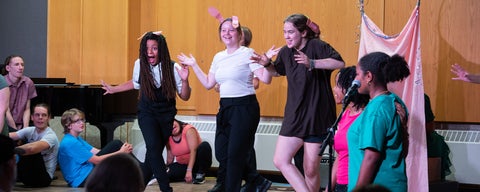 Three young adults dance on stage, wearing paper puppy ears as part of a musical performance.