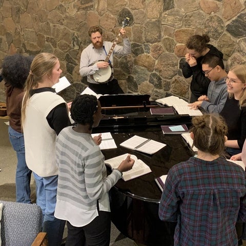 Brandon playing banjo infront of students gathered around a piano