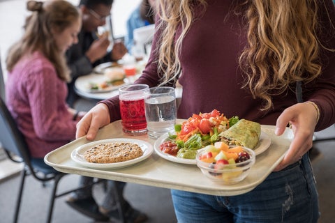 a student holds a tray with plates of salad, a big grebel cookie, and juice
