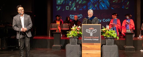 A graduating student stands up at a microphone on stage, while the dean smiles behind a podium to his left. 