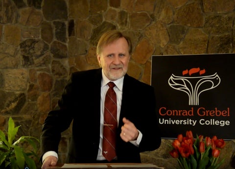 Ed Janzen speaking at podium in front of a sign with Conrad Grebels logo 