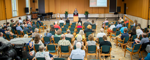 a large room, the great hall, is full of people listening to a speaker on stage