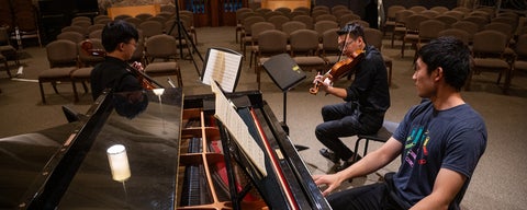 A trio of students, practices in the chapel before a concert. One plays piano, the other a violin, the last a cello. 