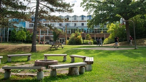 A scenic view of the fire pit outside Grebel, surrounded by green grass. Large trees in the near distance frame the peaked windows of the college residence. A student plays basketball on the court nearby. 