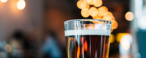 a glass of amber beer sits on a wooden bar, with softly glowing warm lights in the background of a bar