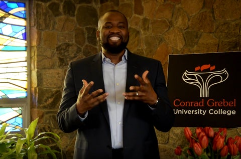 Paul Okoye speaking at podium in front of a sign with Conrad Grebels logo 
