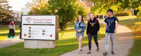 three students walk down a grebel path outside in fall, by the Grebel sign