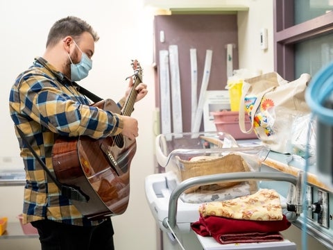 Tyler, wearing a blue mask, plays guitar thoughtfully in a hospital environment, specifically to an unseen baby in a medical basinet.