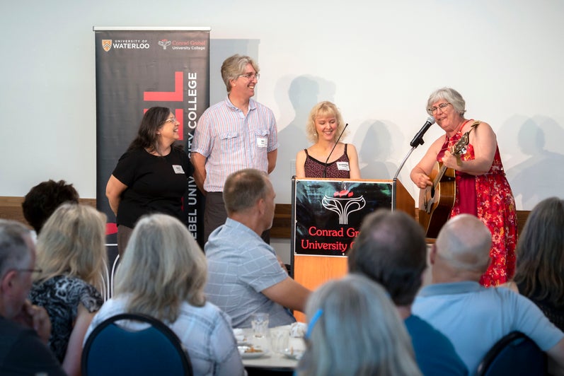 Four Grebel alumni stand at the front of the room, leading the crowd in a song, accompanied by a guitar.