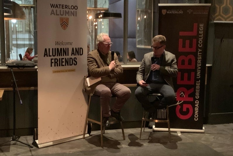 Peter Harder and Marcus Shantz sit and discuss in front of a University of Waterloo and Grebel banner.