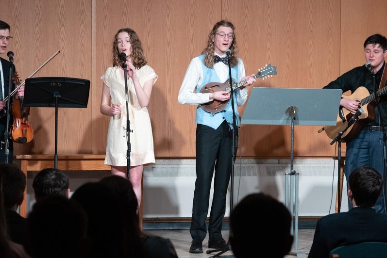 four students singing and playing instruments in front of a crowd 
