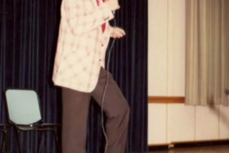 a student stands at a microphone, performing some sort of story (80s)