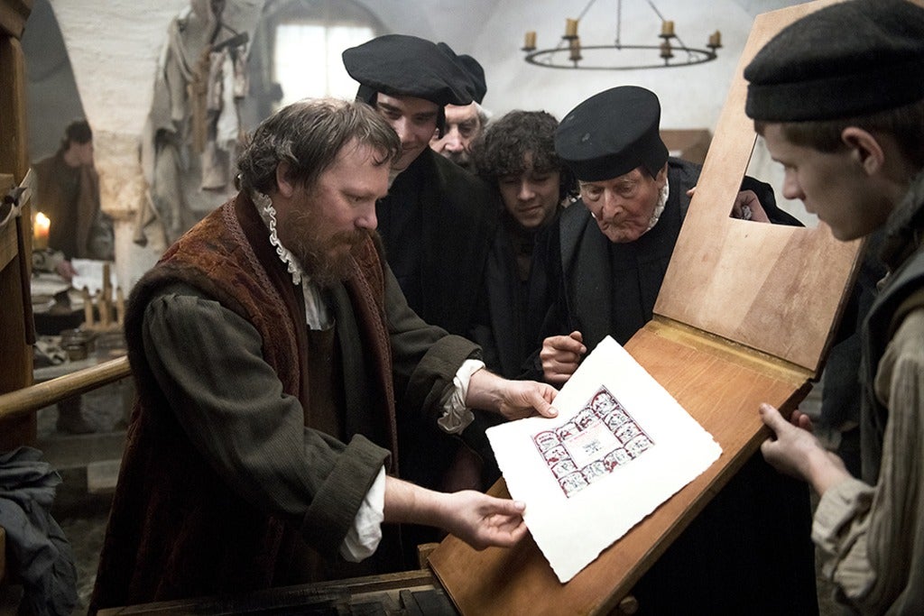 A scene, where a group of men dressed in medieval european clothes, all gather around a board with an illuminated piece of paper. One of the men holds it gently, while the others look on.