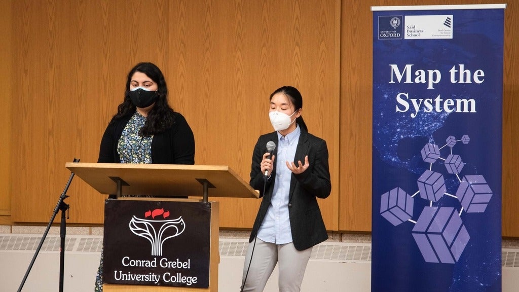 two students standing at a podium weairng masks beside a banner displaying the words "map the system"