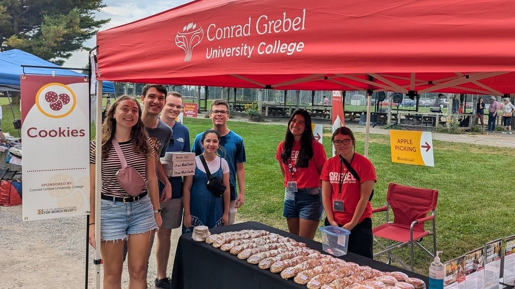 Similing Grebel students stand under a red tent at a park