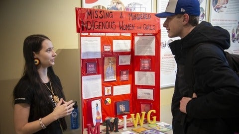 two students talking while stanidng in fornt of a poster board with information on missing and murdered indigenous women.