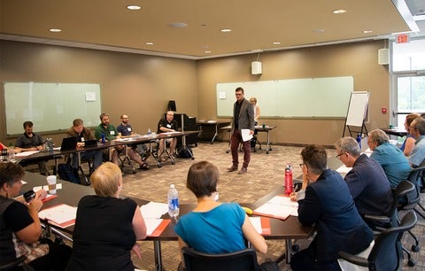 group of people sat around a horseshoe table with someone standing in the middle speaking 