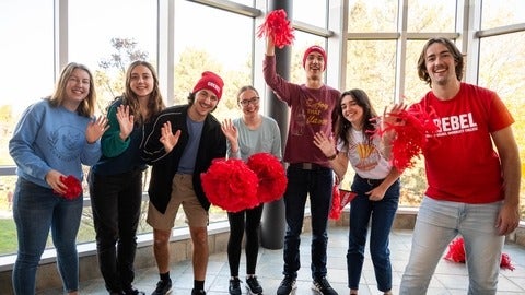 A group of seven Grebel students with red pompoms smile and wave.