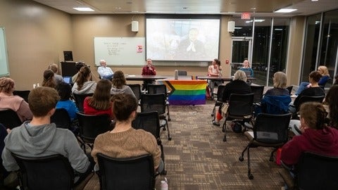 people sitting in chairs facing a table at the front of the room with a pride flag hanging from it