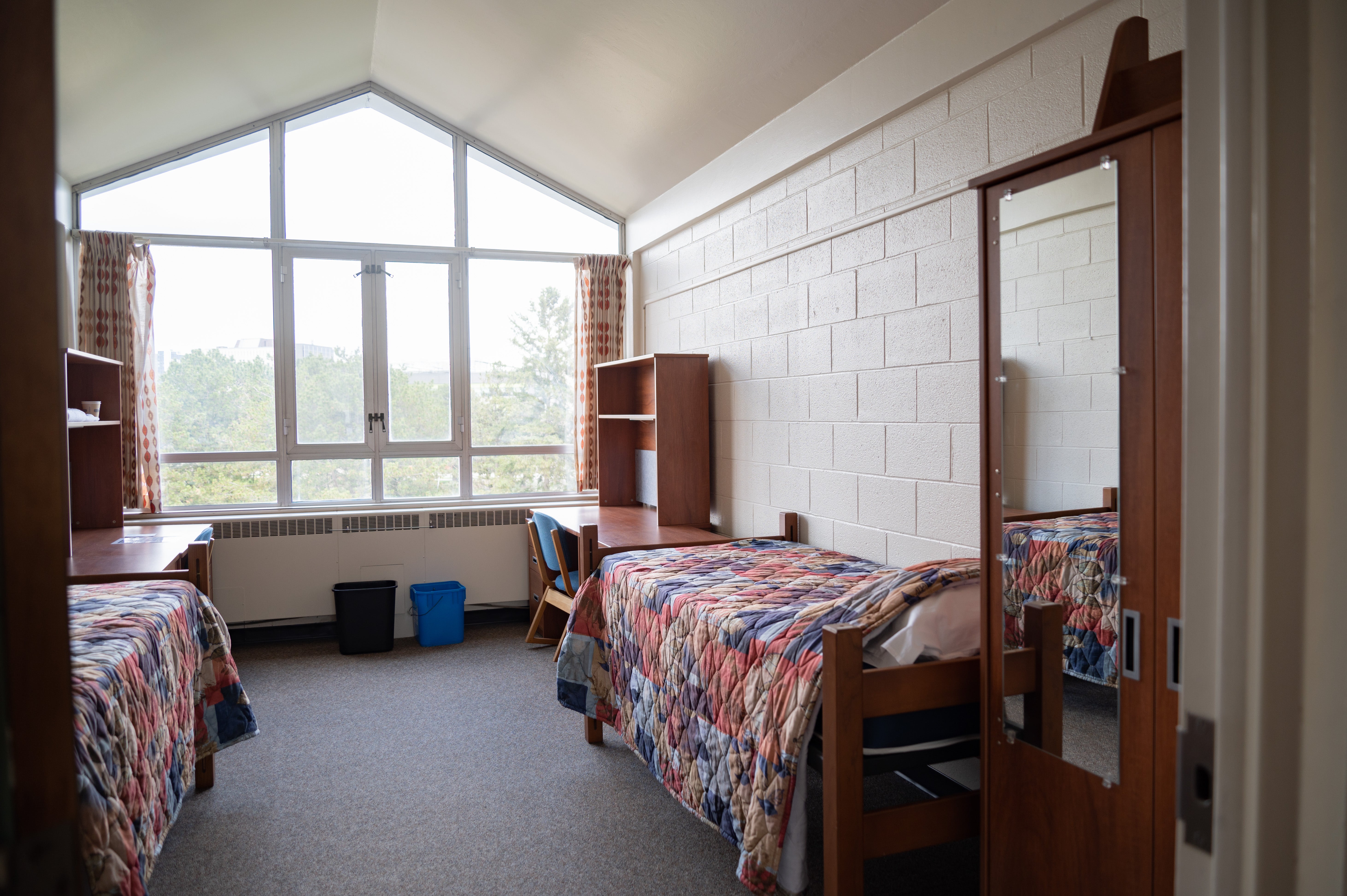 a room with two beds with colourful quilts, and two desks. A large window that ends in a peak above lets light into the room.