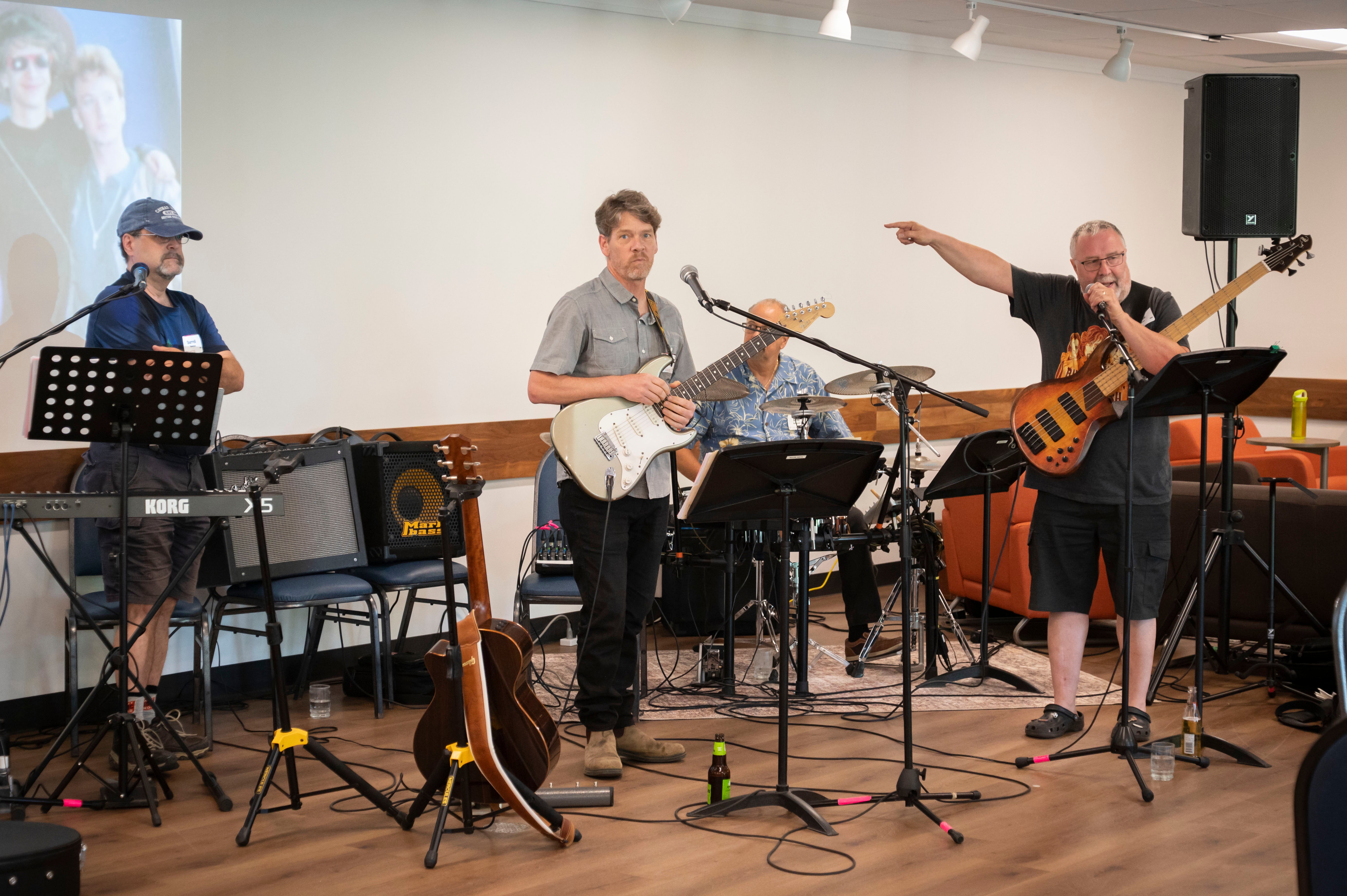 A band of four Grebel Alumni. One points to a screen. They have guitars, and a drum kit, and a piano.